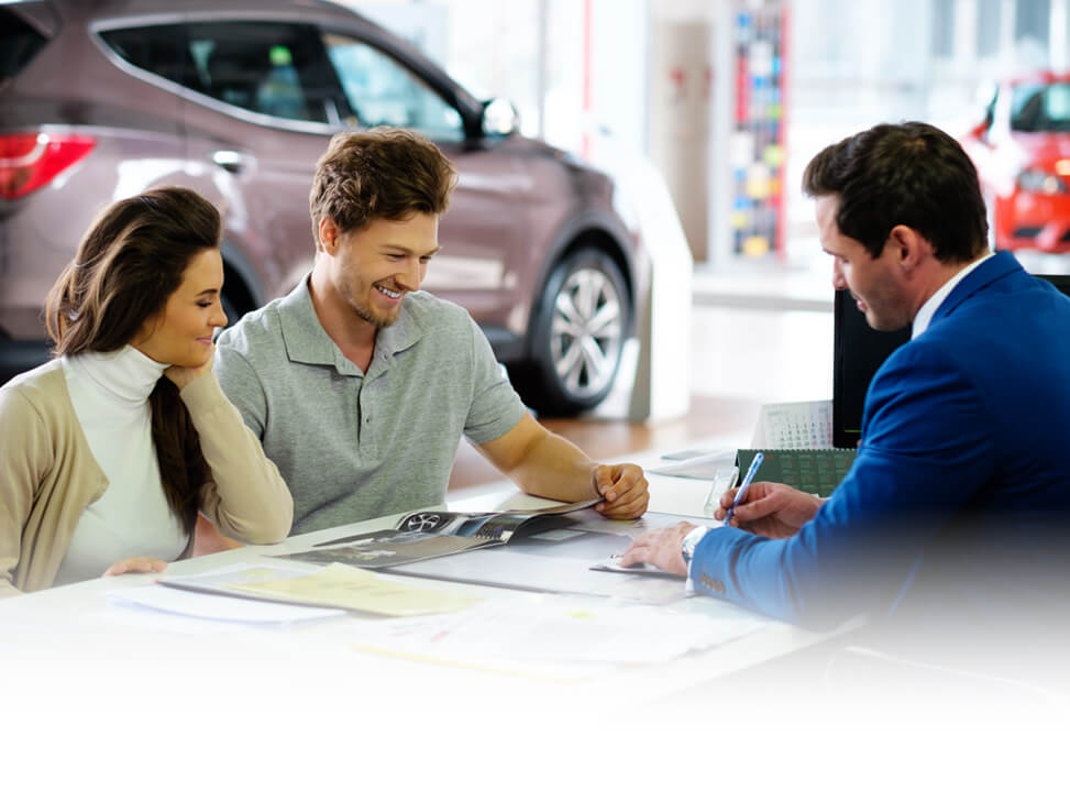 Man And Woman At Car Dealership Going Over Paperwork With Salesman